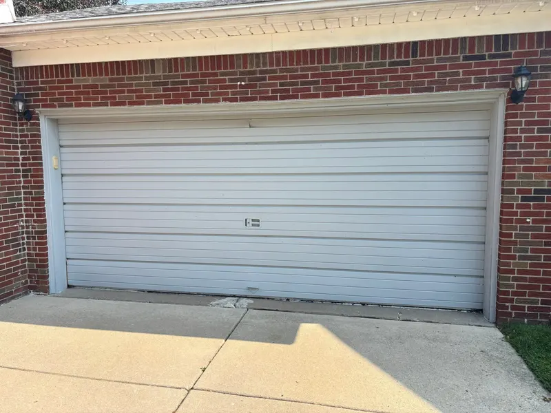 Old white double garage door with a dented top section and a worn bottom seal, on a brick house